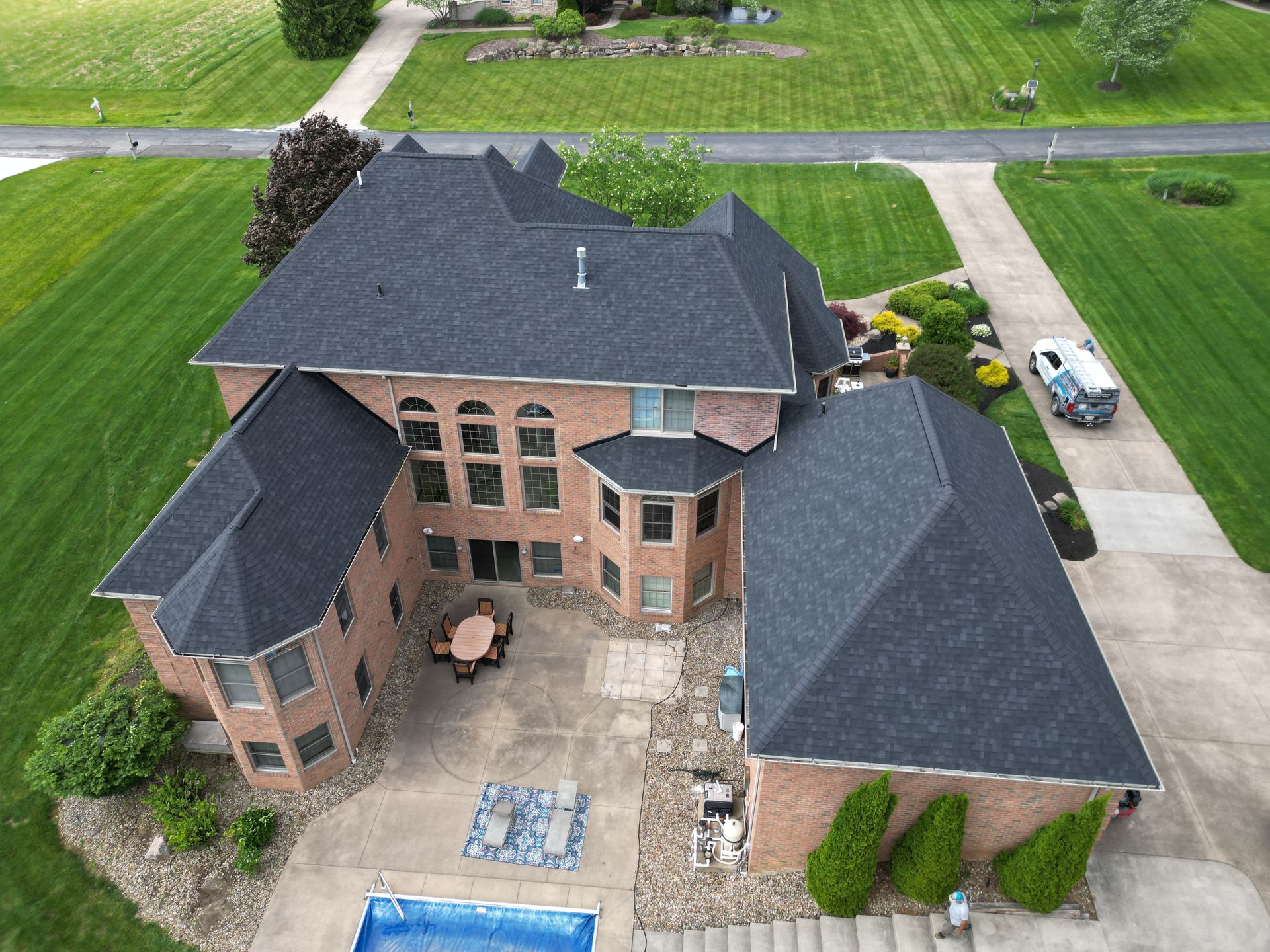 Aerial view of a large brick house with a dark gray roof, patio, pool, and a long driveway.