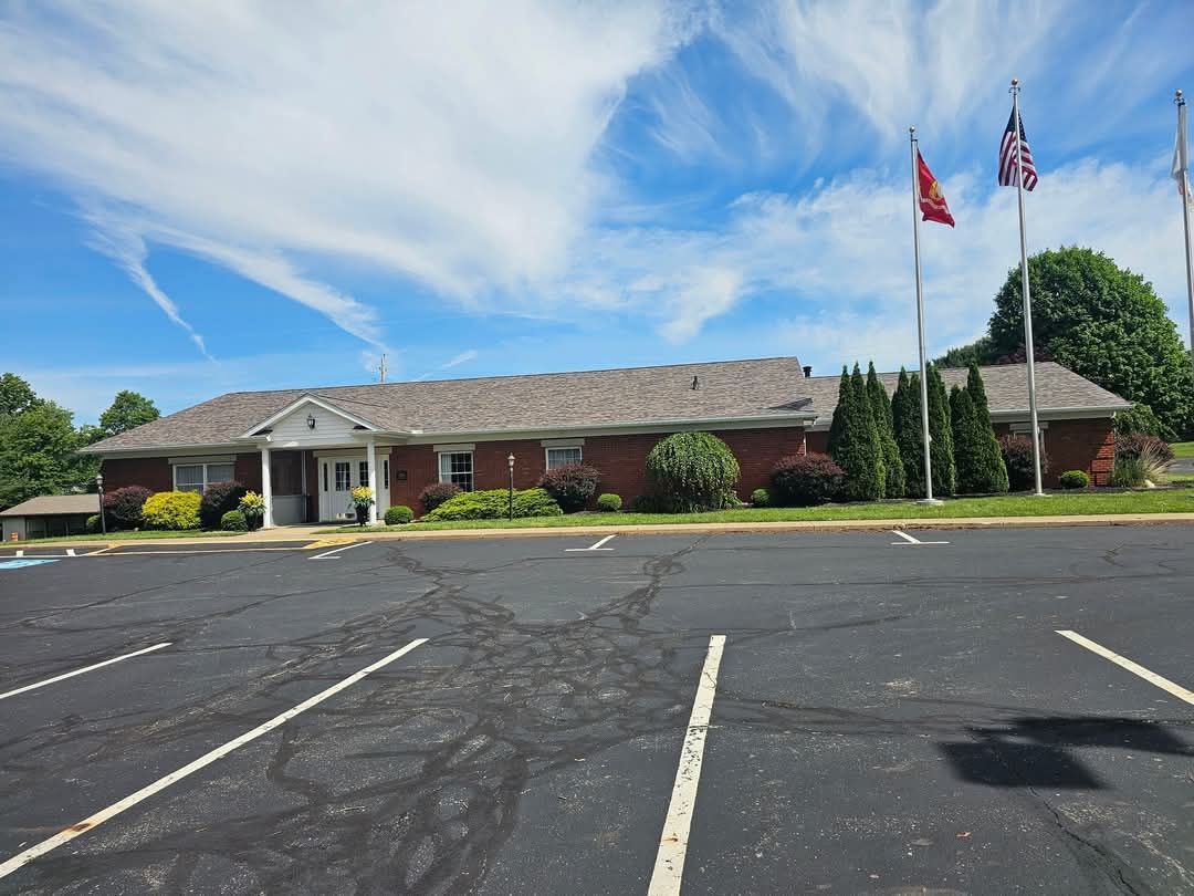 A one-story brick building with flags in front, a large parking lot, and a partly cloudy blue sky.