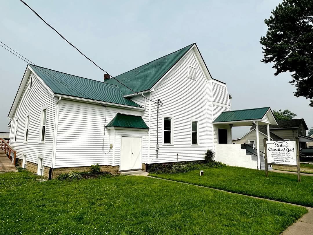 White building with green roof and trim, likely a church, with a grassy lawn and sign.