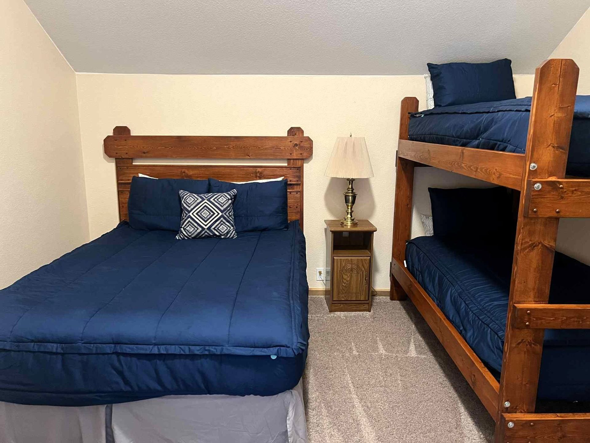 Small bedroom with blue bedding, a queen bed, and a wooden bunk bed beside a nightstand lamp.
