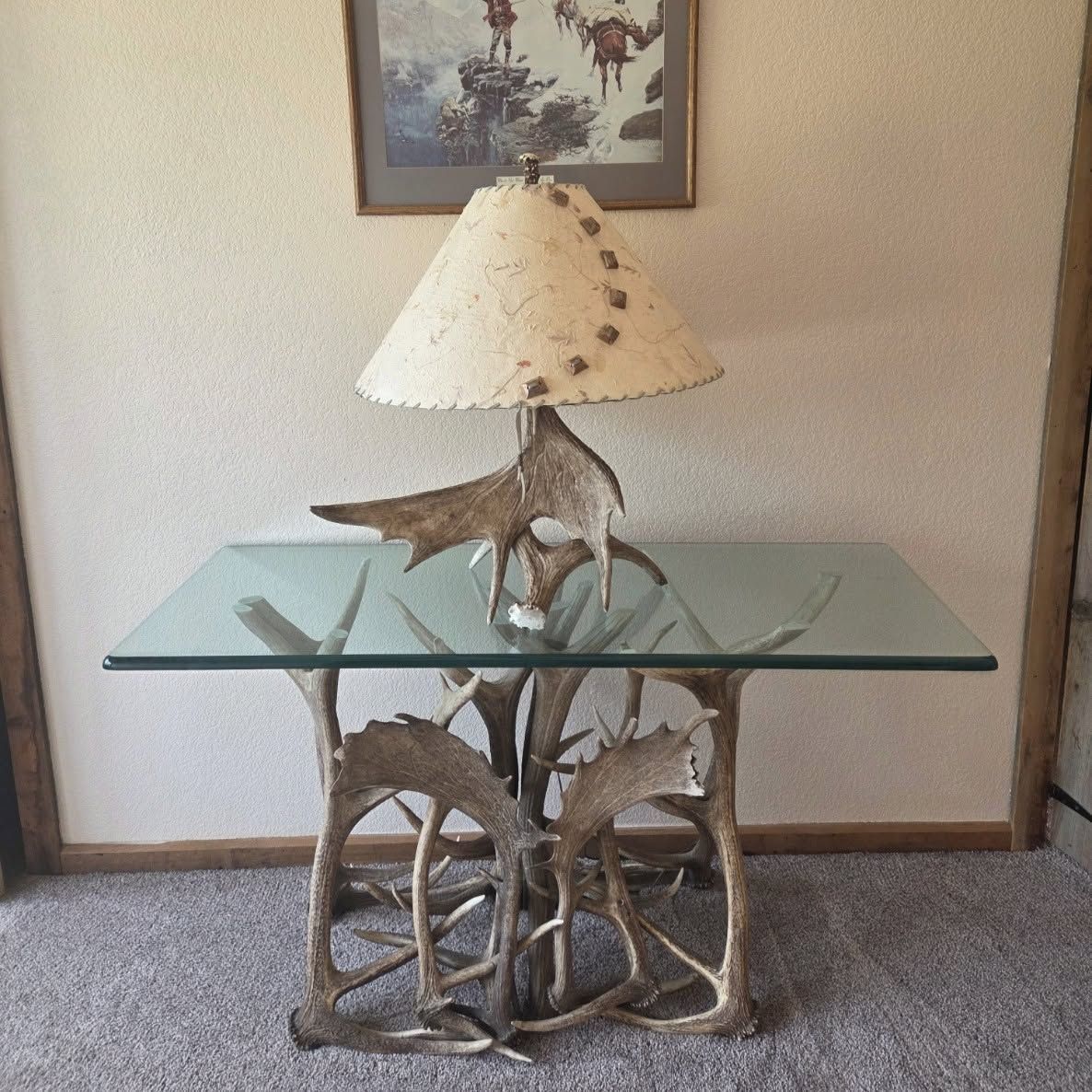 Glass-top table with sculptural metal base and conical lamp shade in a carpeted room