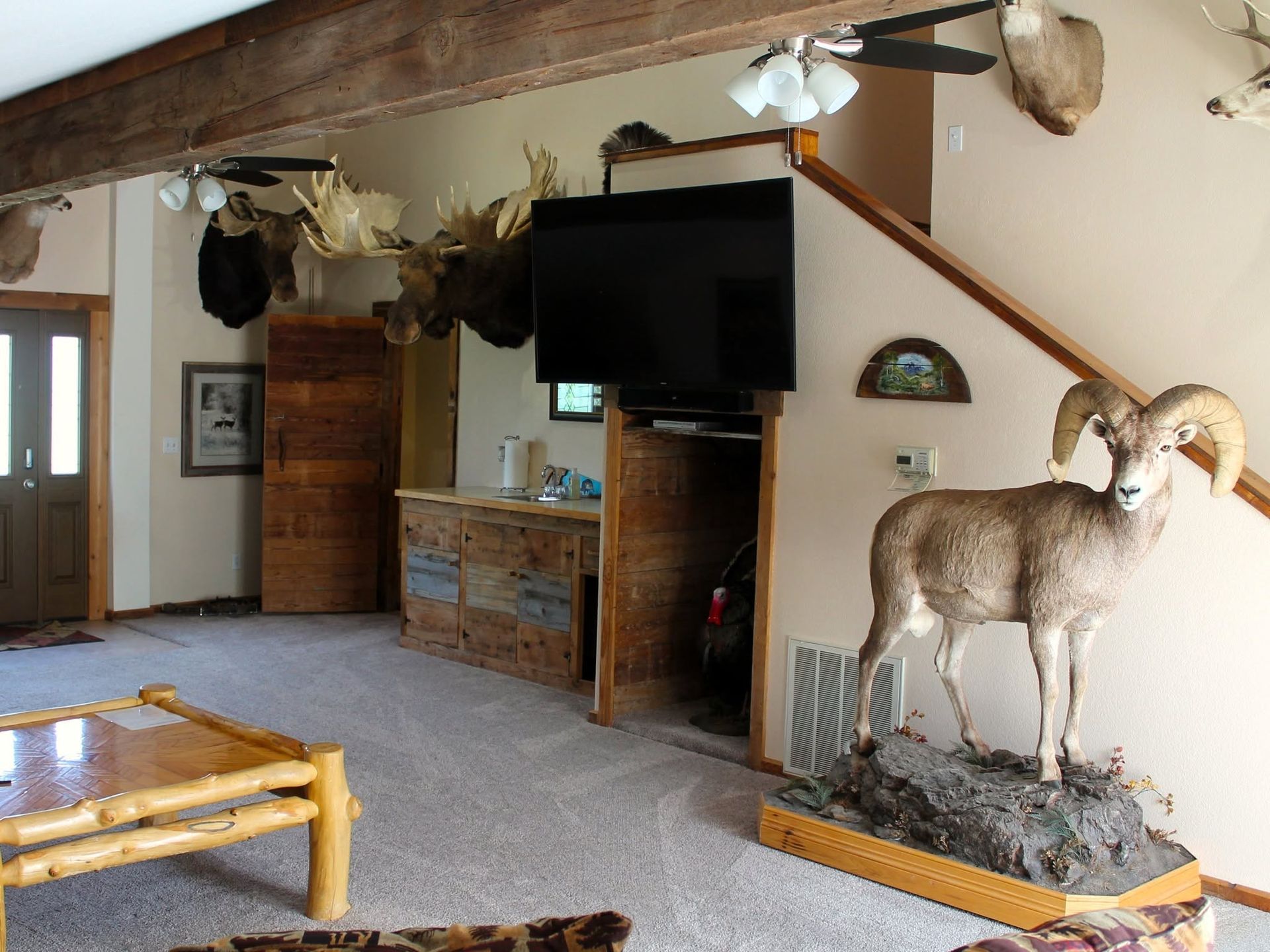 Rustic living room with mounted TV, wooden furniture, and a ram taxidermy display on a stone base.