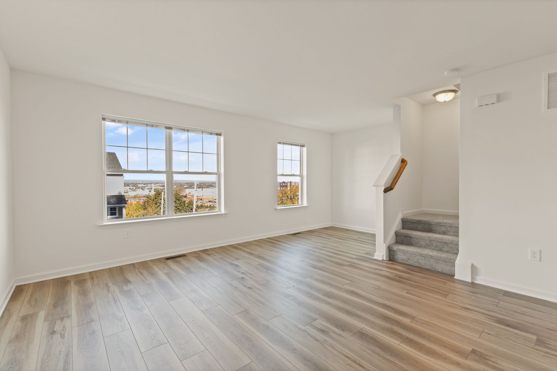 Empty bright living room with wood floors, large windows, and carpeted stairs.