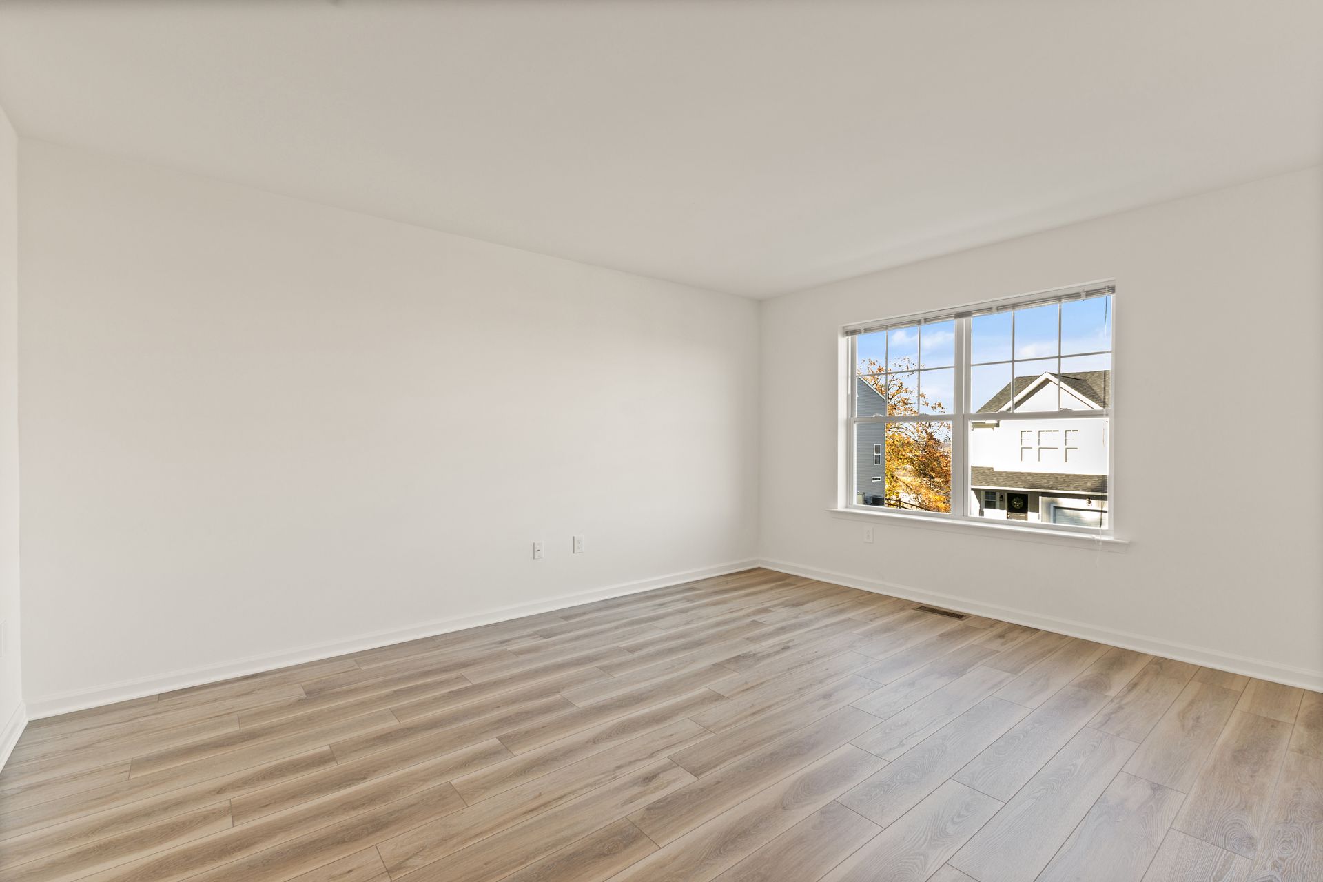 Empty white bedroom with light wood floors and a window overlooking houses and autumn trees