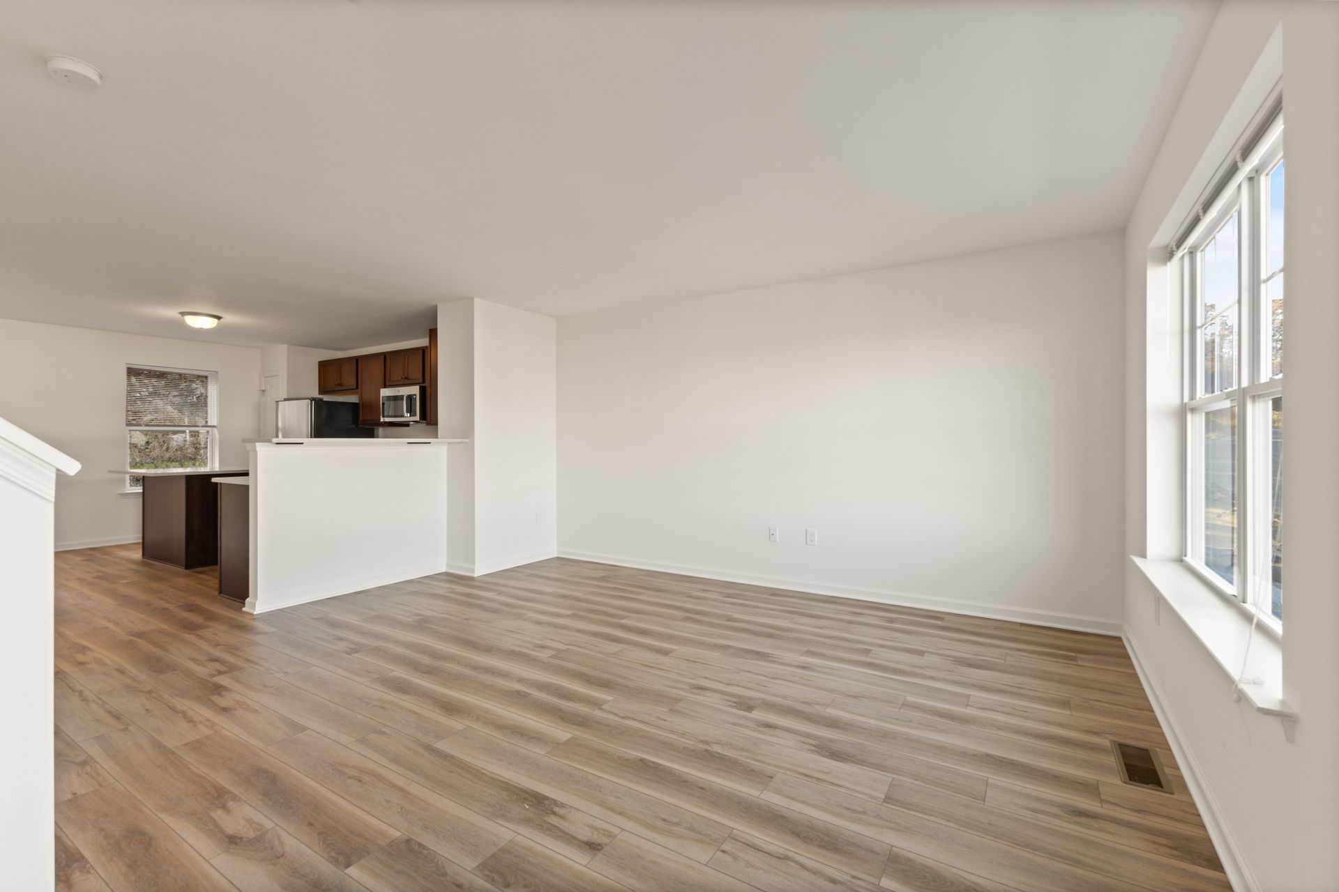 Empty white living room with wood flooring and a kitchen nook in the back left