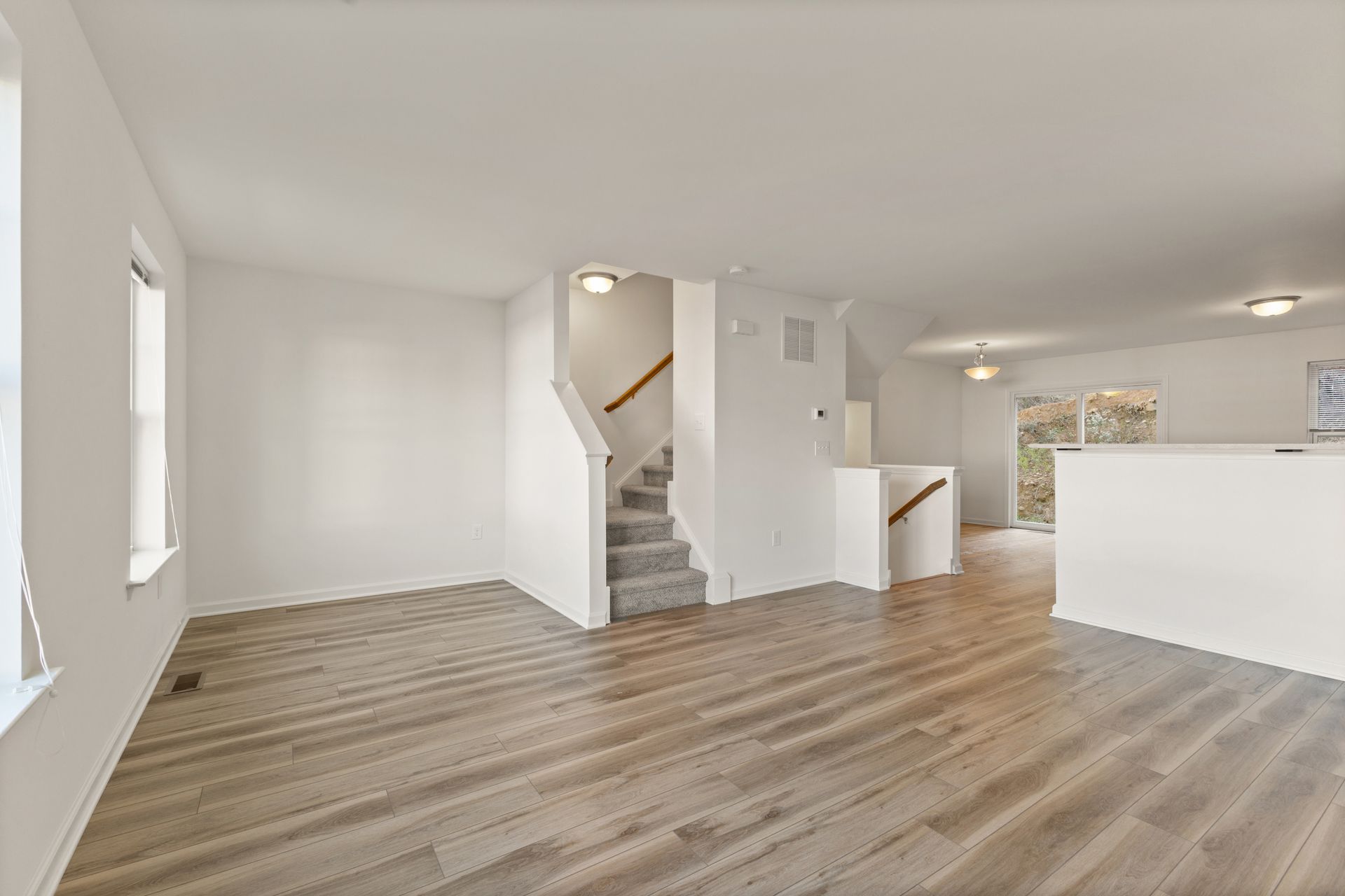 Empty modern living room with wood flooring, white walls, staircase, and open kitchen area