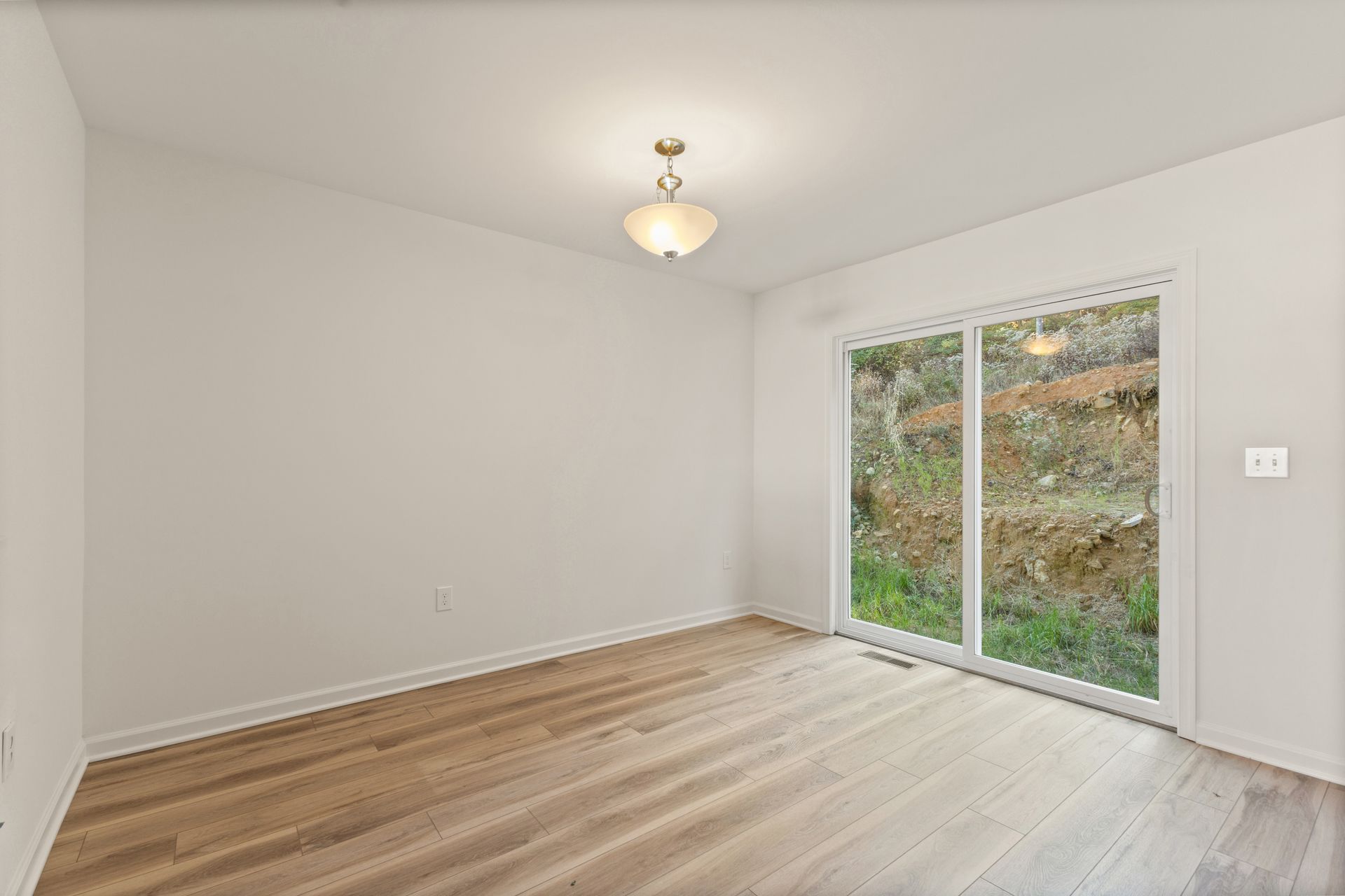 Empty white room with wood floors and sliding glass door to a leafy outdoor area