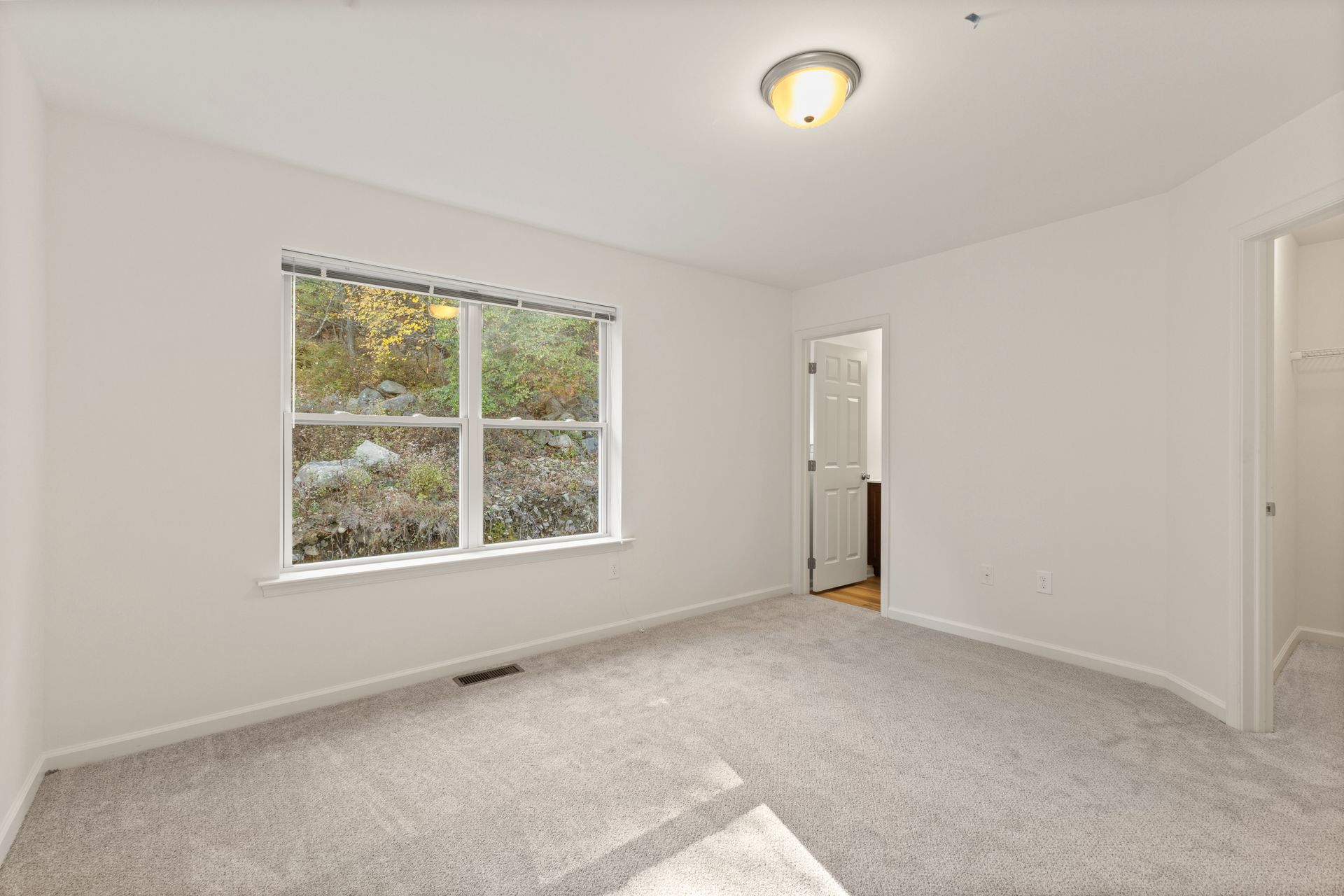 Empty carpeted bedroom with white walls, window, ceiling light, and open doorway