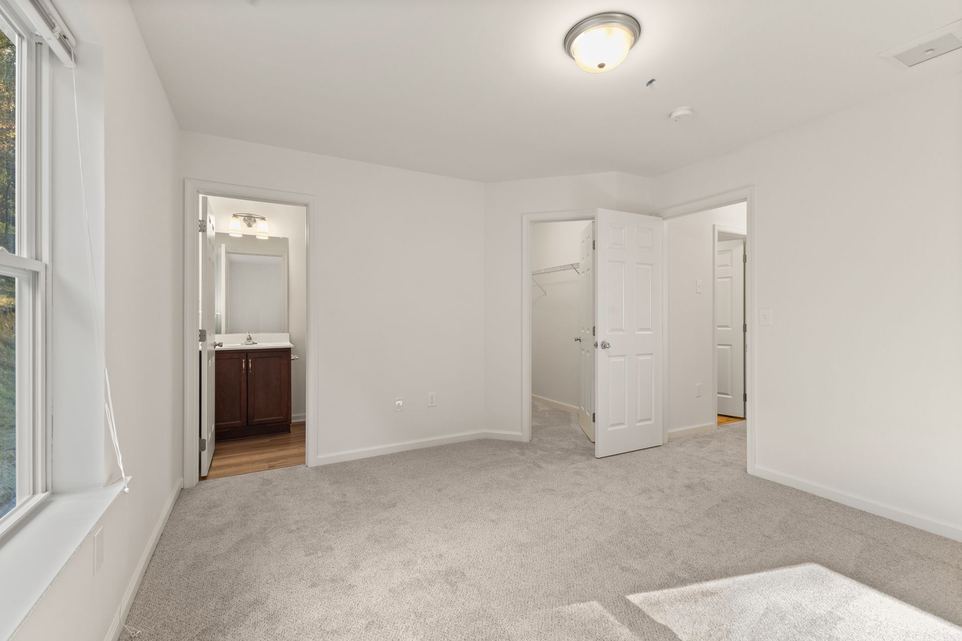Empty carpeted bedroom with white walls, window seat, closet doors, and adjoining bathroom vanity visible