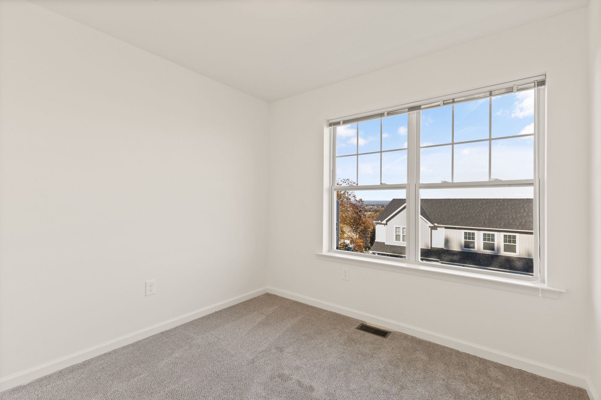 Empty white bedroom with gray carpet and a window overlooking neighboring houses