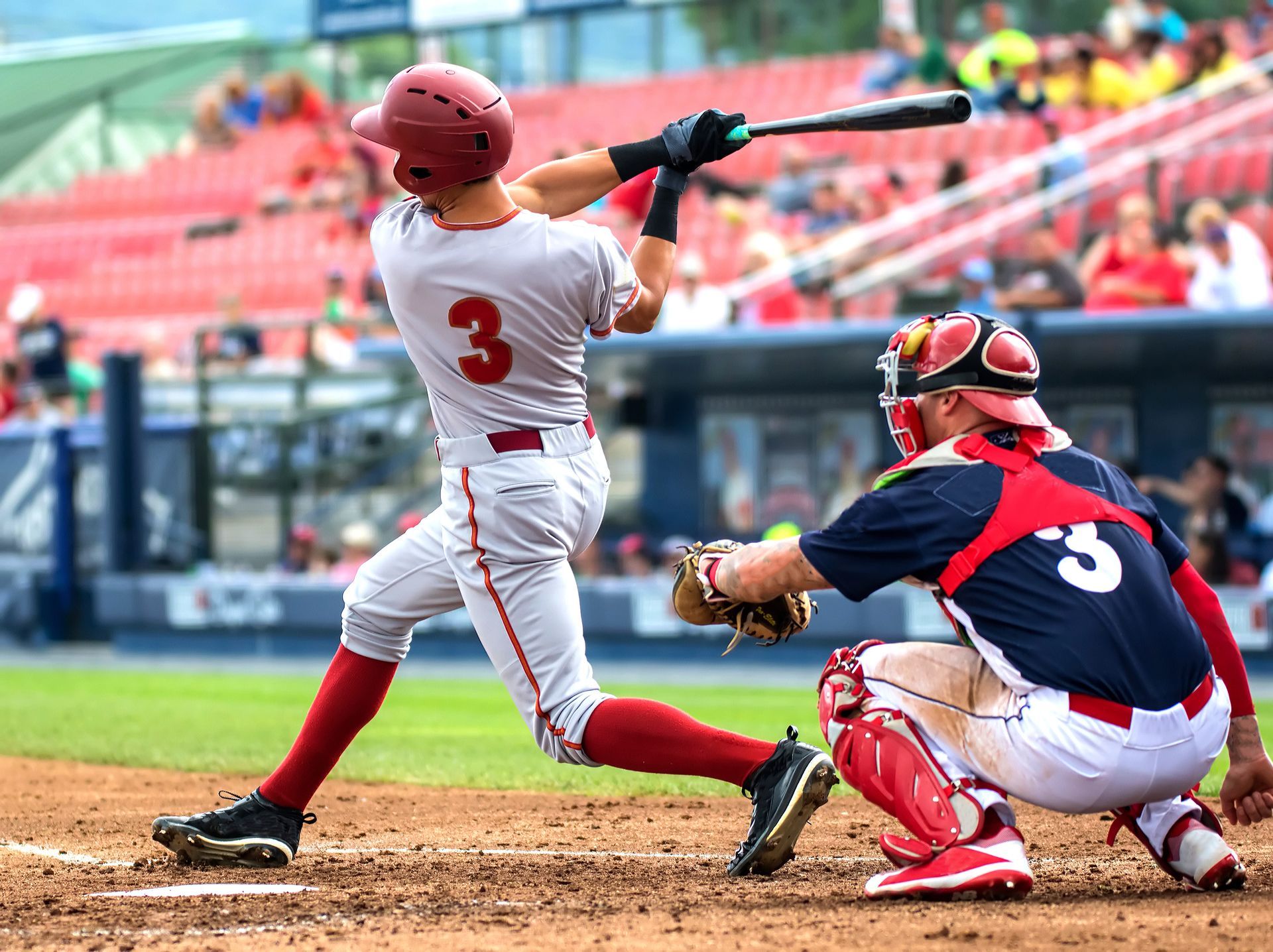 Baseball batter in red swings at pitch as catcher in navy crouches behind home plate