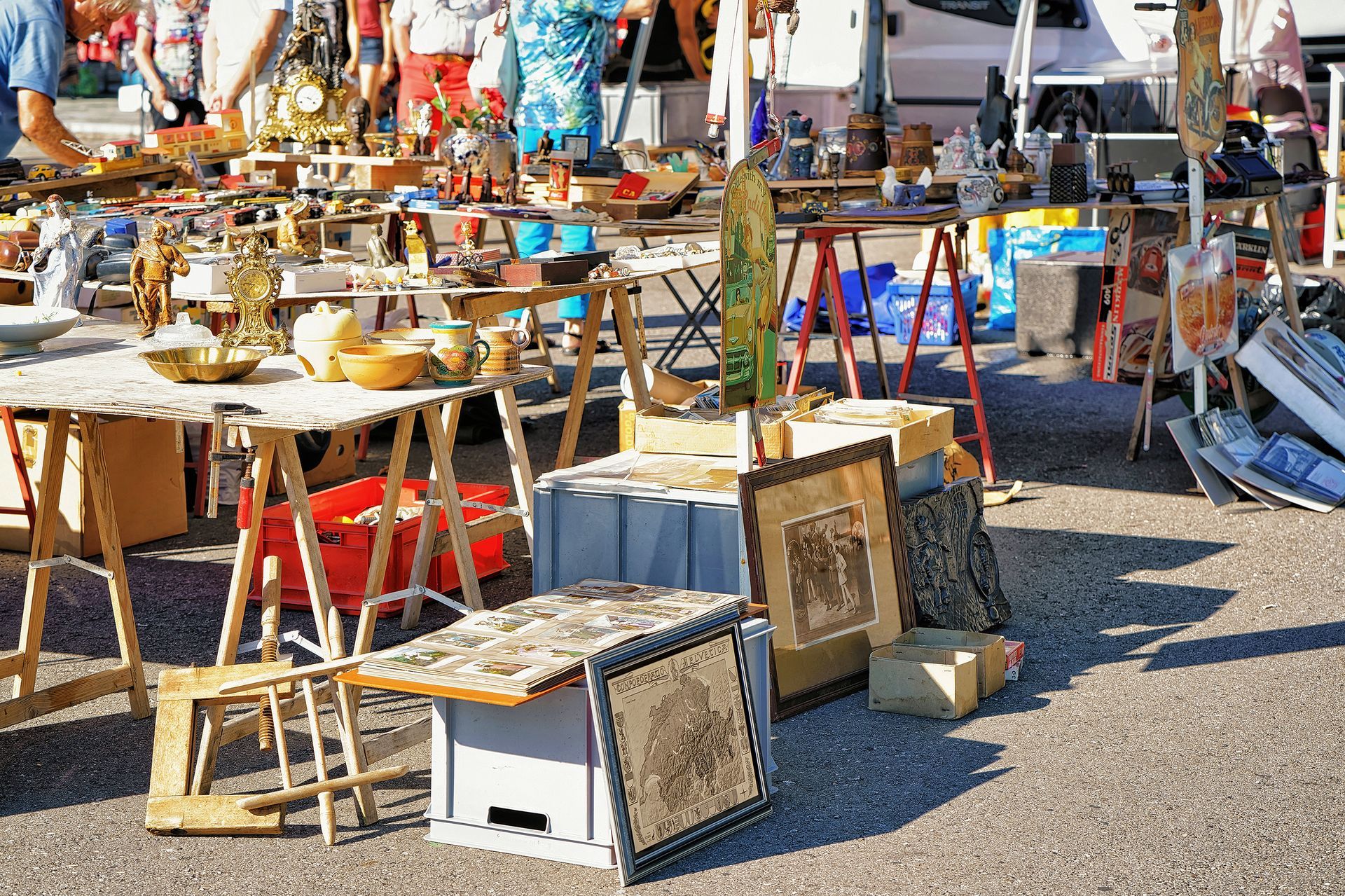 Outdoor flea market with tables of assorted items and framed artwork displayed on gravel.
