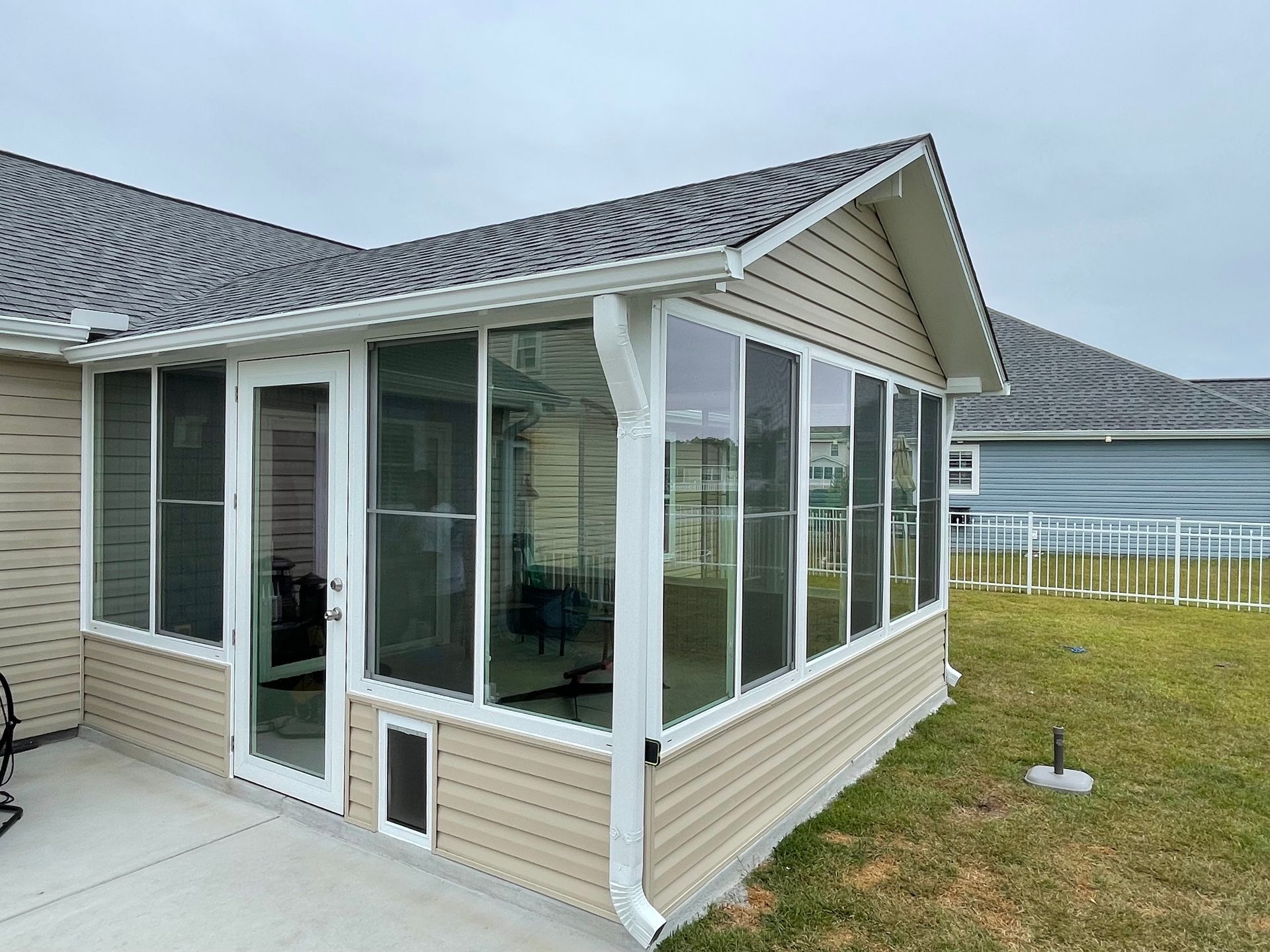 Screened-in porch addition with white frames and tan siding, next to a house with dark gray roof, on a lawn.
