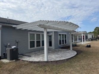 White pergola over a curved patio attached to a gray house; cloudy sky in background.