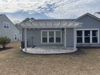 A backyard patio with a pergola, gray siding, and a stone patio. Overcast sky.