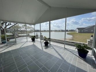 Screened porch overlooking a lake, featuring gray tiled floor, potted plants, and a bright, sunny view.