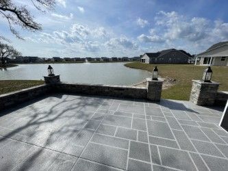 Stone patio overlooking a lake, with decorative lanterns on pillars. Houses in the distance under a cloudy sky.