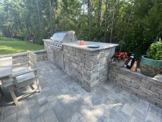 Outdoor kitchen with grill, counter, and patio seating. Gray brick construction with surrounding greenery.