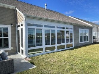 A sunroom with white frames and many windows, attached to a gray house. Green grass in the foreground.