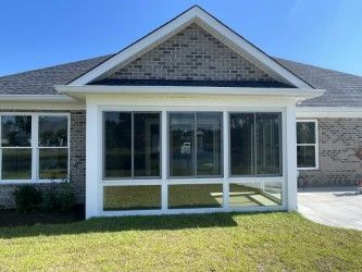 A sunroom addition with glass windows, white trim, and a gray brick exterior. It is on a grassy lawn with a blue sky.
