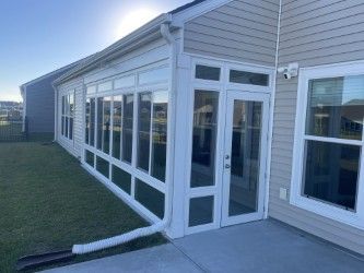 Sunroom with white trim and glass windows attached to a beige house, next to green grass and a concrete patio.