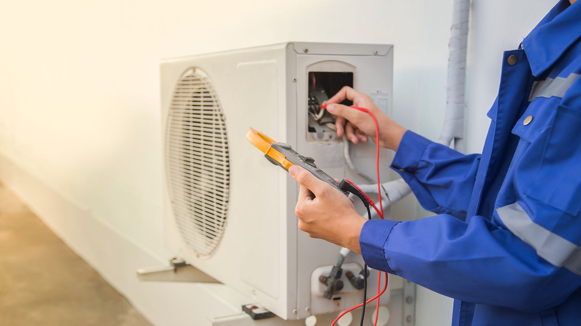 HVAC technician in blue uniform testing an outdoor air conditioning unit with a multimeter.