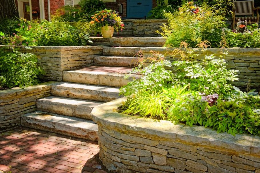 Stone steps with tiered landscaping leading to a blue door. Brick and lush greenery frame the steps.