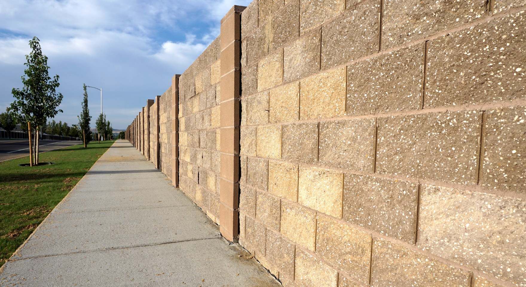 Tall tan block wall running beside a sidewalk and grassy area for property support during a block and retaining wall service in Santa Ana, CA