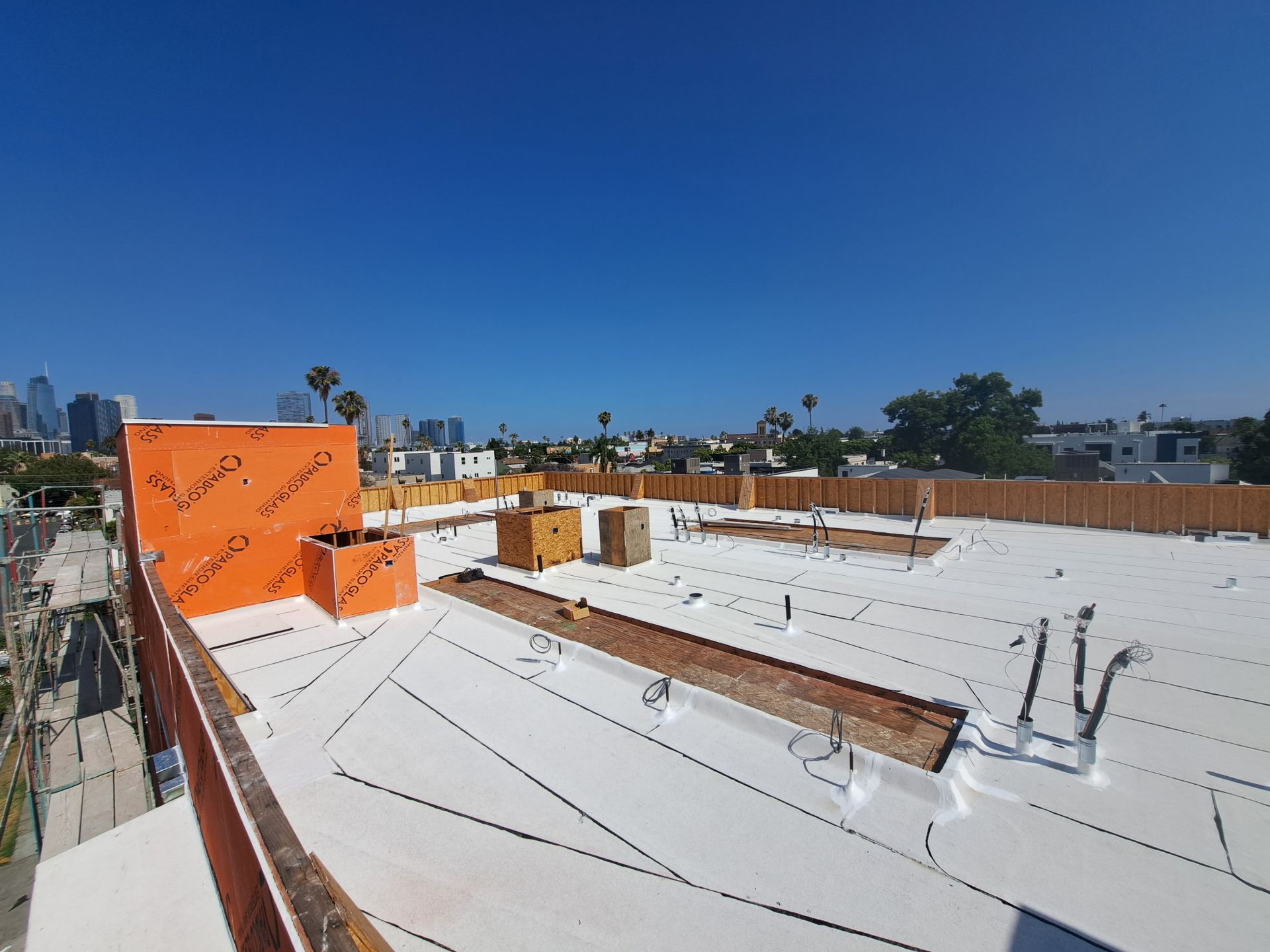 Rooftop under construction with white waterproofing, orange tarp, and city skyline under a clear blue sky