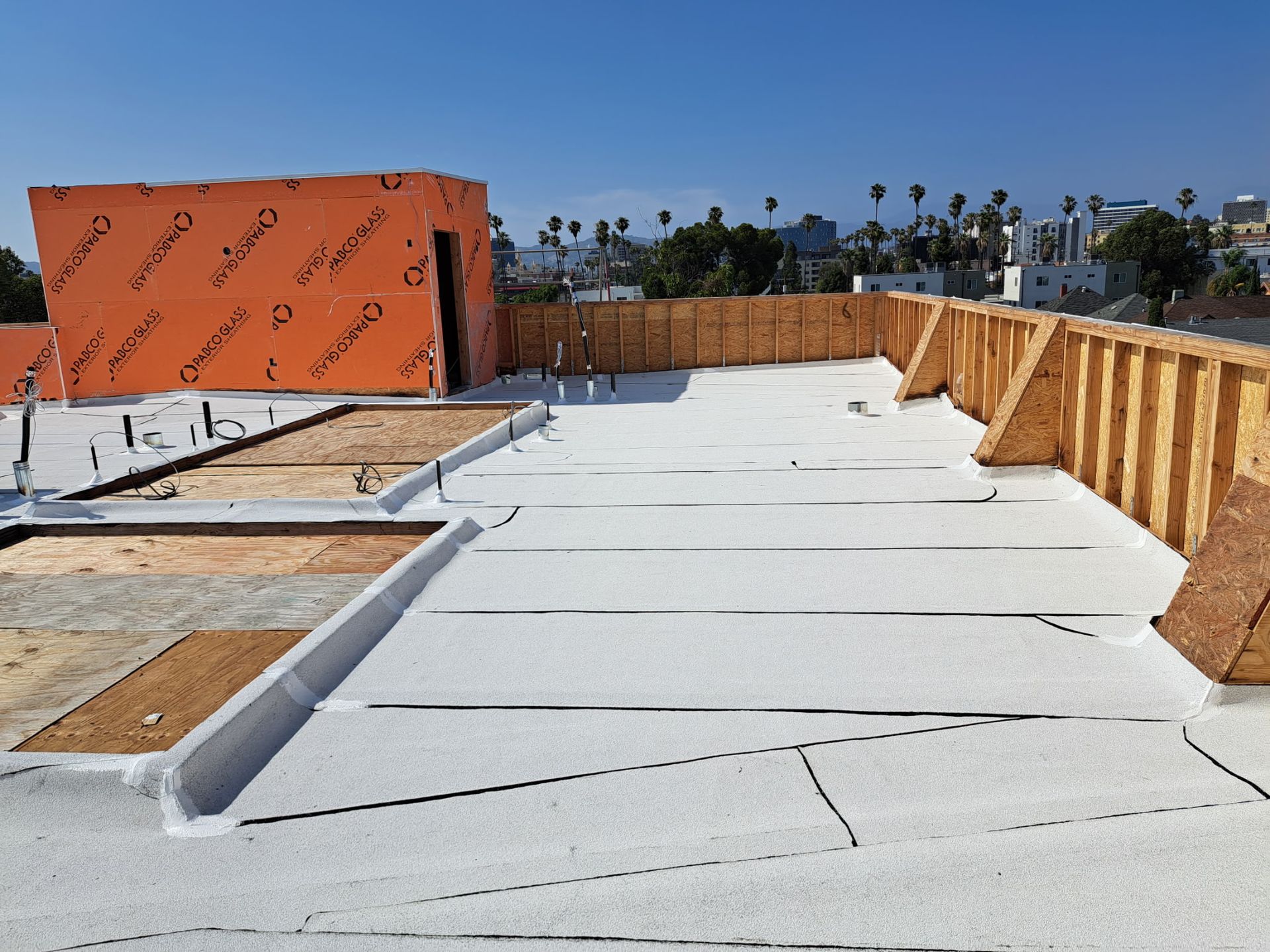 Flat rooftop under construction with white roofing, orange sheathing, and wooden framing against a blue sky