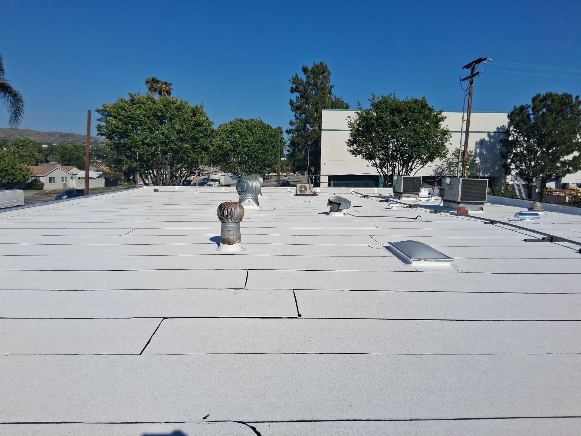 Flat white rooftop with vents and skylights, trees and buildings visible under a clear blue sky.