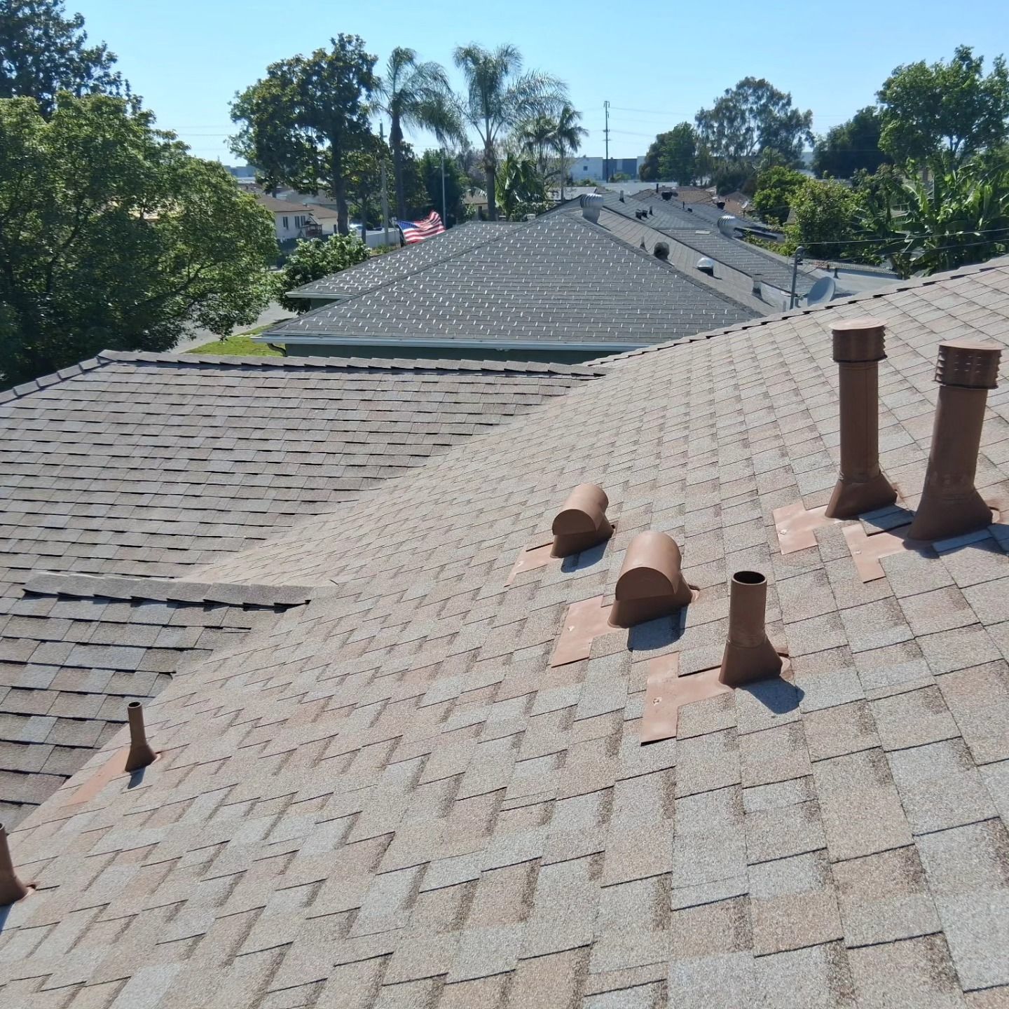 Rooftop with gray shingles and several vent pipes overlooking trees and neighboring houses on a sunny day