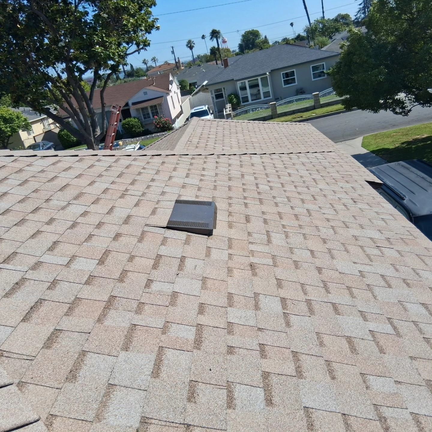 Rooftop with gray shingles and a small roof vent, overlooking a sunny suburban neighborhood.