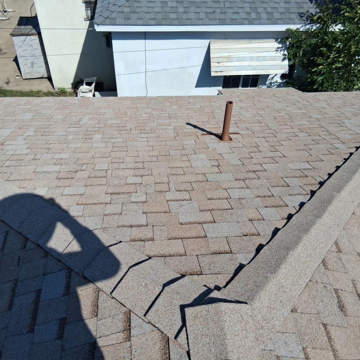 Roof shingles with a protruding vent pipe and visible cracks near the ridge in daylight