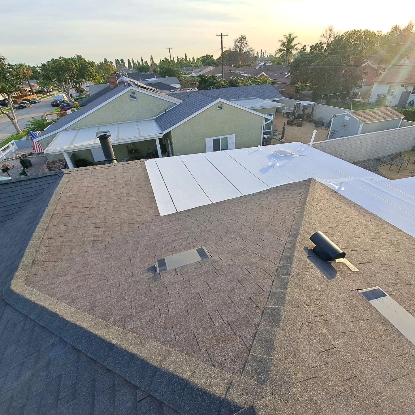 Rooftop view of suburban houses at sunset, with white roof panels and a distant street lined with trees.
