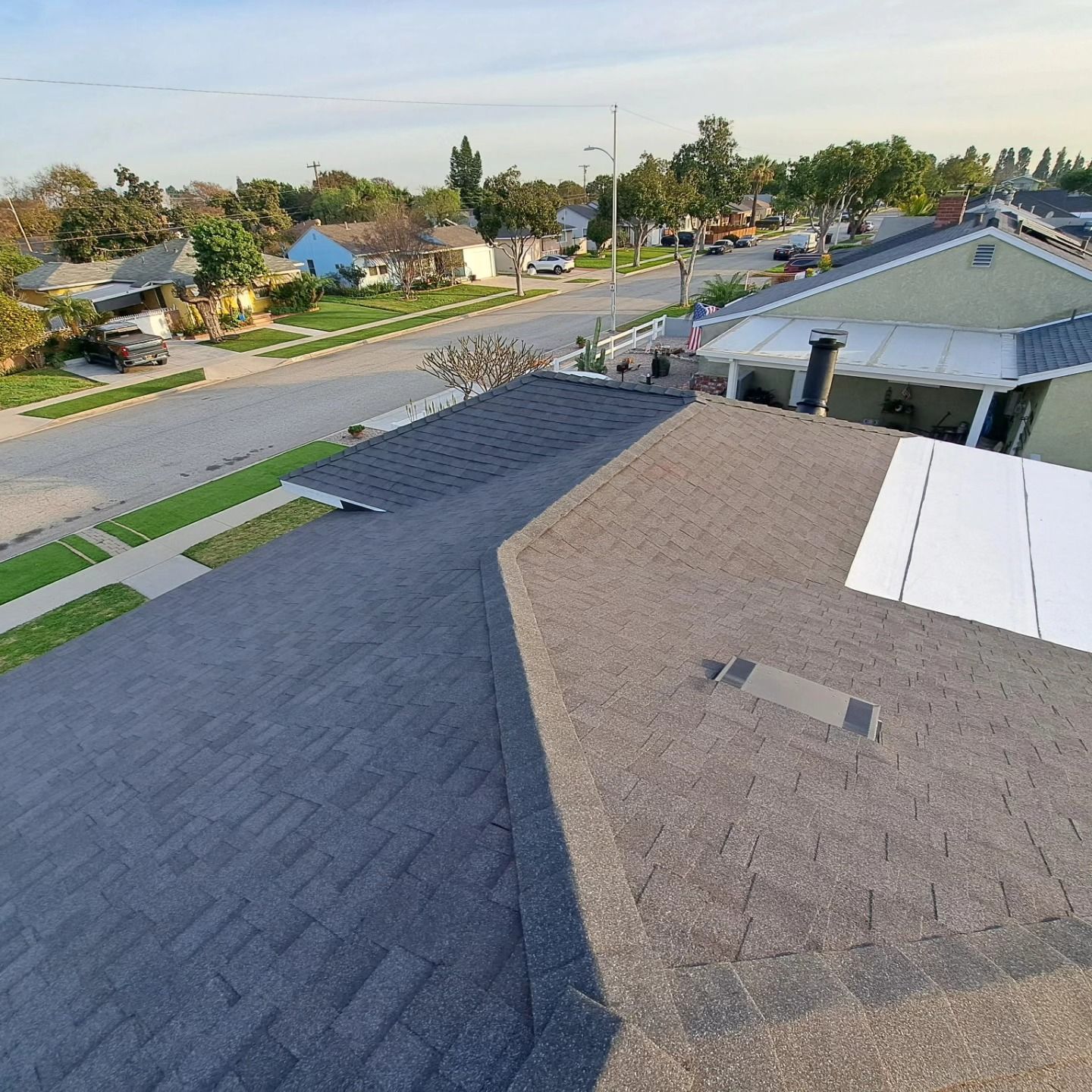 Rooftop view of a suburban street with houses, trees, and a partially shingled roof in the foreground