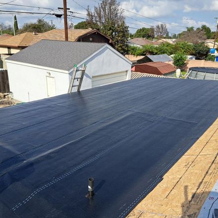 Flat dark-blue roof being installed on a house, with a ladder and neighborhood homes in the background.