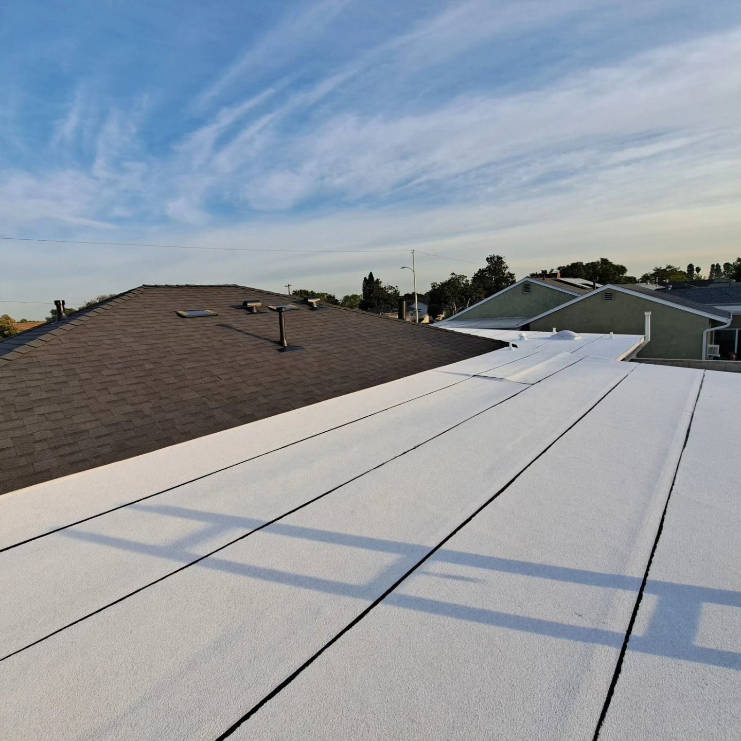 White flat rooftop with seams beside a brown shingled roof under a cloudy sky
