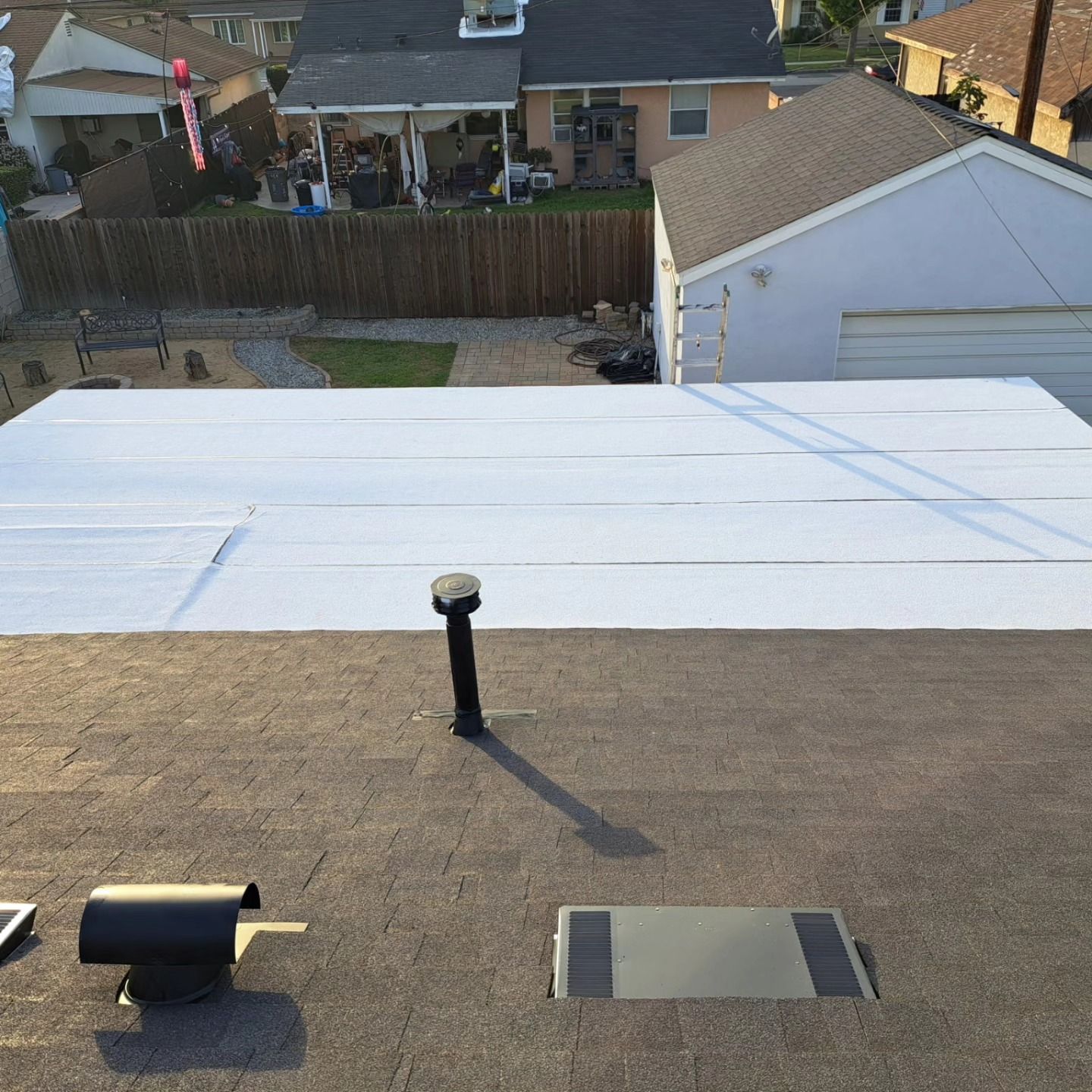 Flat rooftop with a large white tarp, black vent pipes, and neighboring houses in the background.