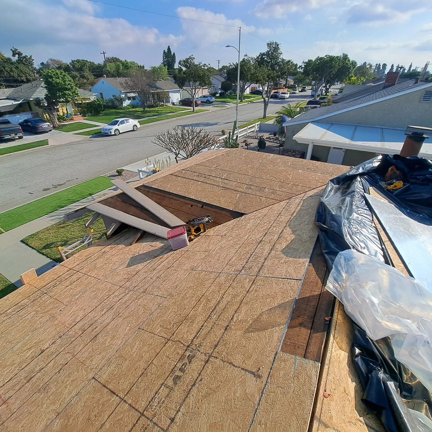 Rooftop under construction with plywood, tarp, and tools, overlooking a quiet suburban street on a sunny day