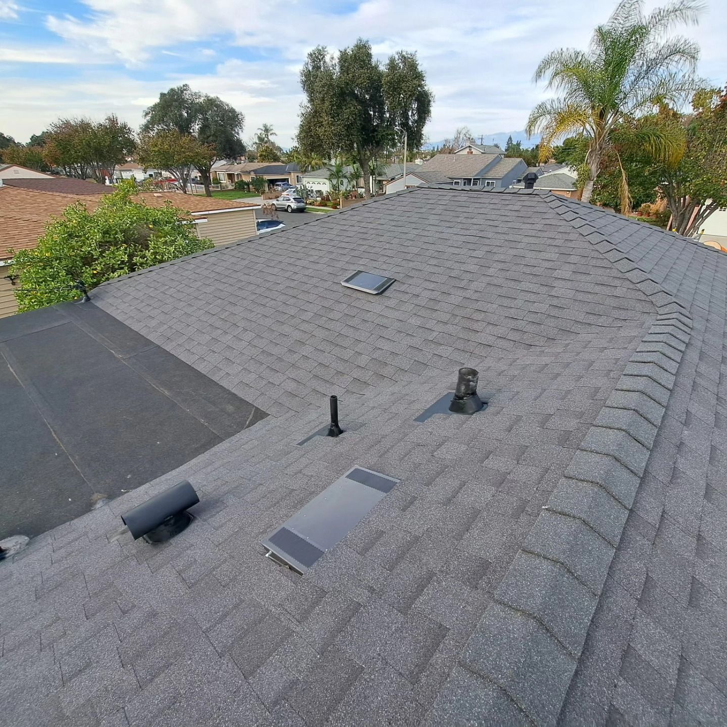 Rooftop with gray shingles, vents, and skylights in a suburban neighborhood under a blue sky