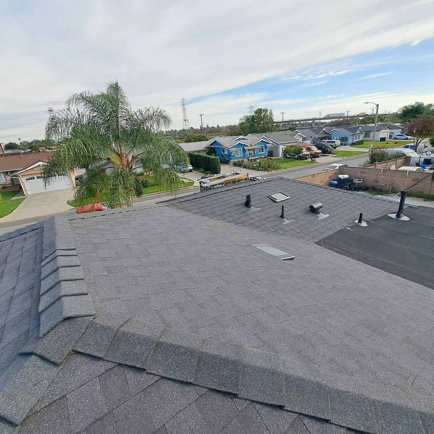 Rooftop shingles and vents overlooking a suburban street with parked cars and trees under a cloudy sky