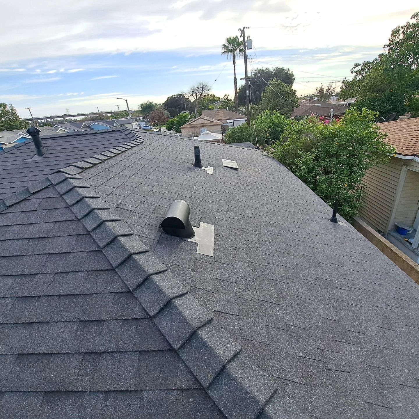 Residential roof with dark shingles and gray gravel, showing rooftop vents and a suburban neighborhood in the background