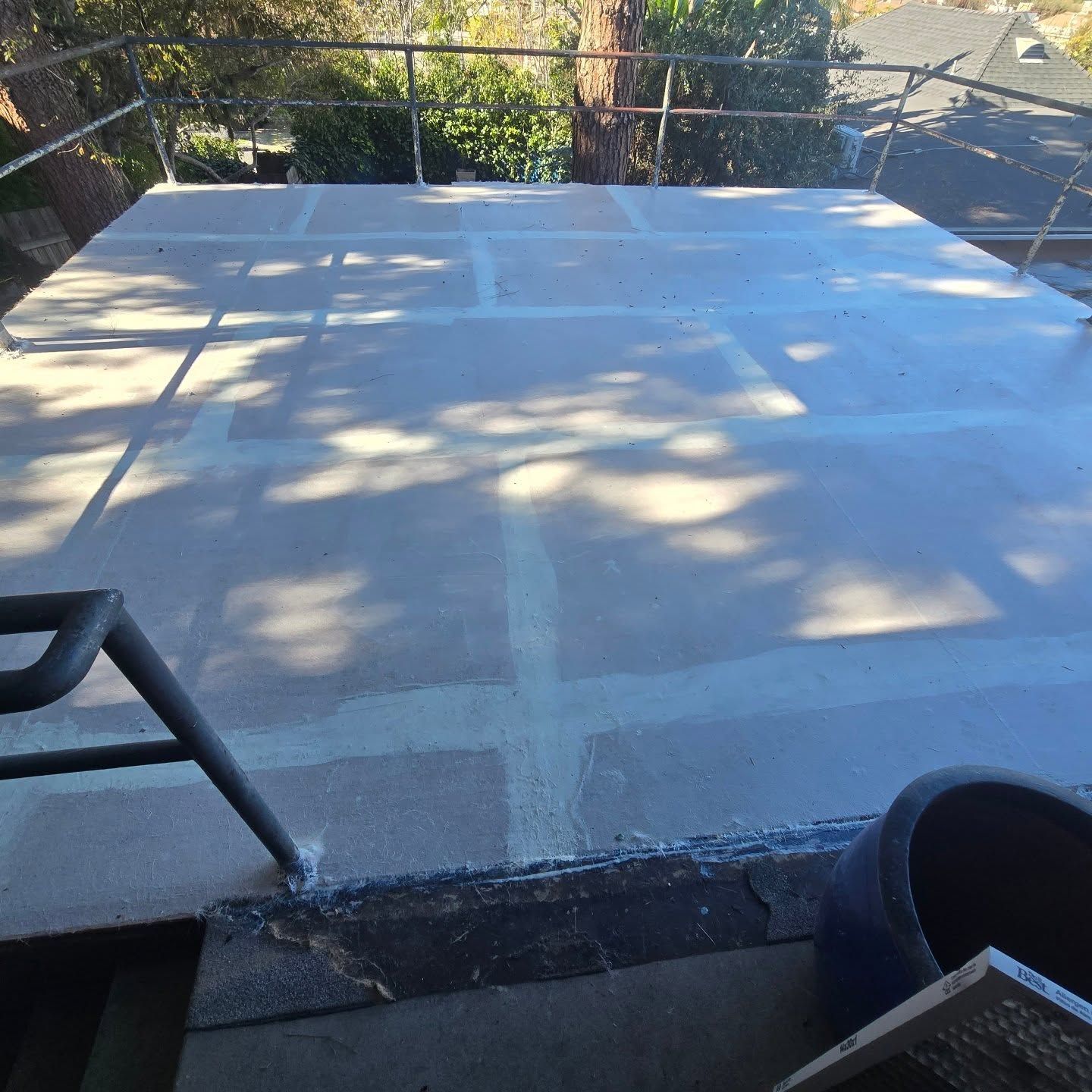 Flat rooftop deck with light gray coating, tree shadows, and a chair and planter in the foreground.