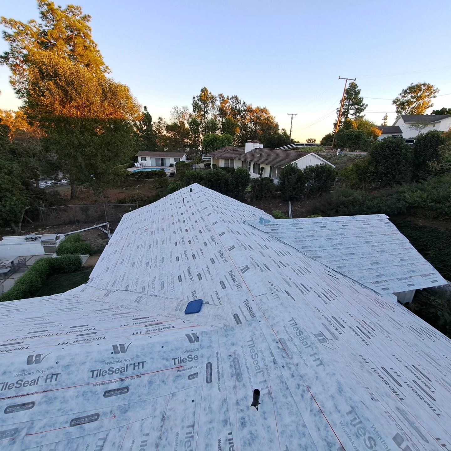 Roof under construction covered with white underlayment at sunset, with trees and houses in the background