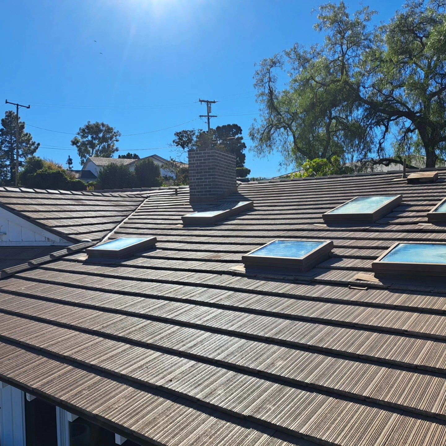 Rooftop with brown shingles, skylight windows, chimney, and trees under a bright blue sky