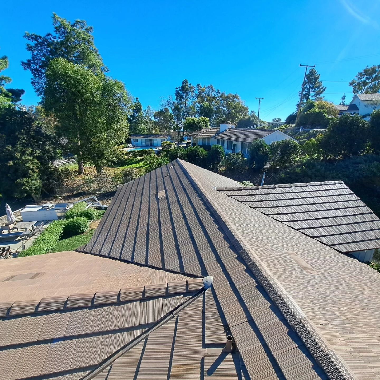 Rooftops with gray shingles beneath a bright blue sky, surrounded by green trees and houses.