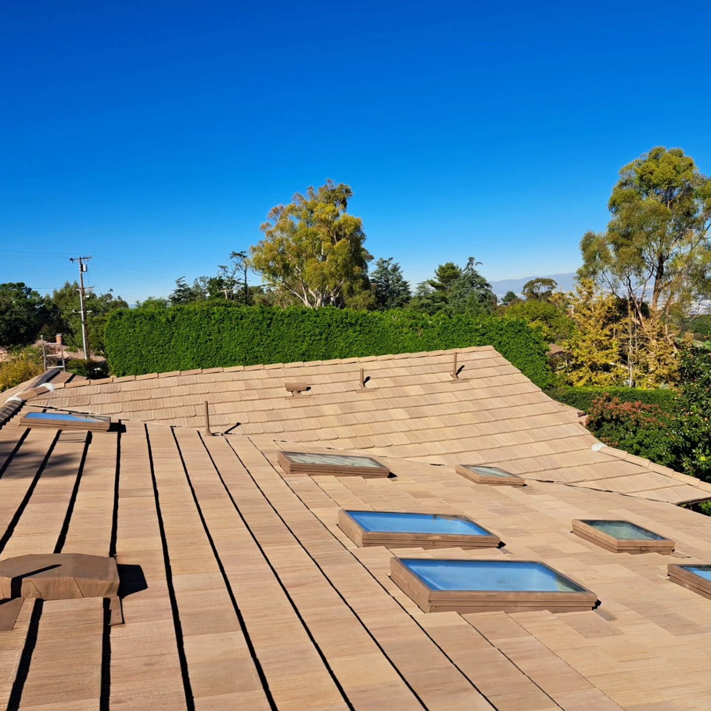 Roof under construction with tan tiles, skylights, and a clear blue sky behind trees