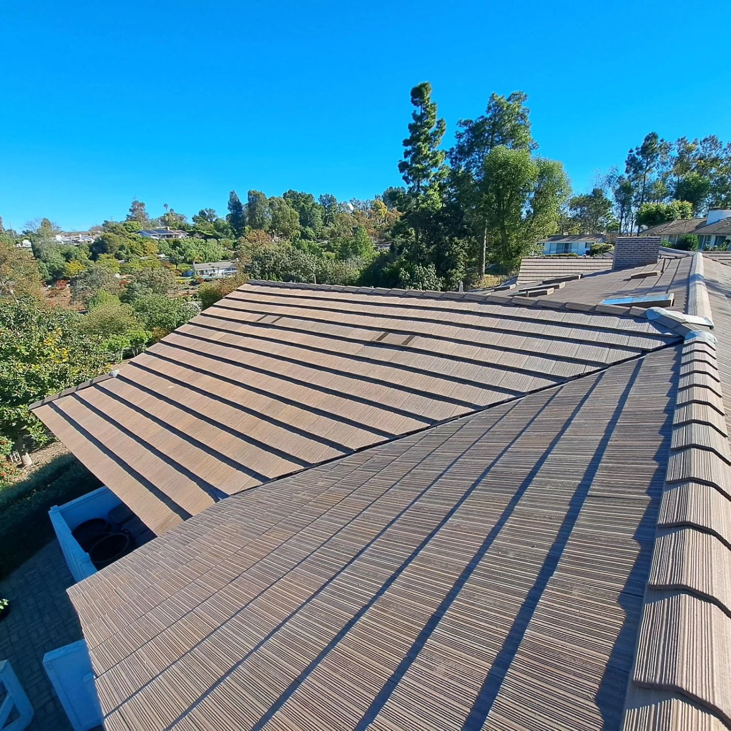 Residential roof with brown shingles under a clear blue sky, surrounded by trees and hillside homes