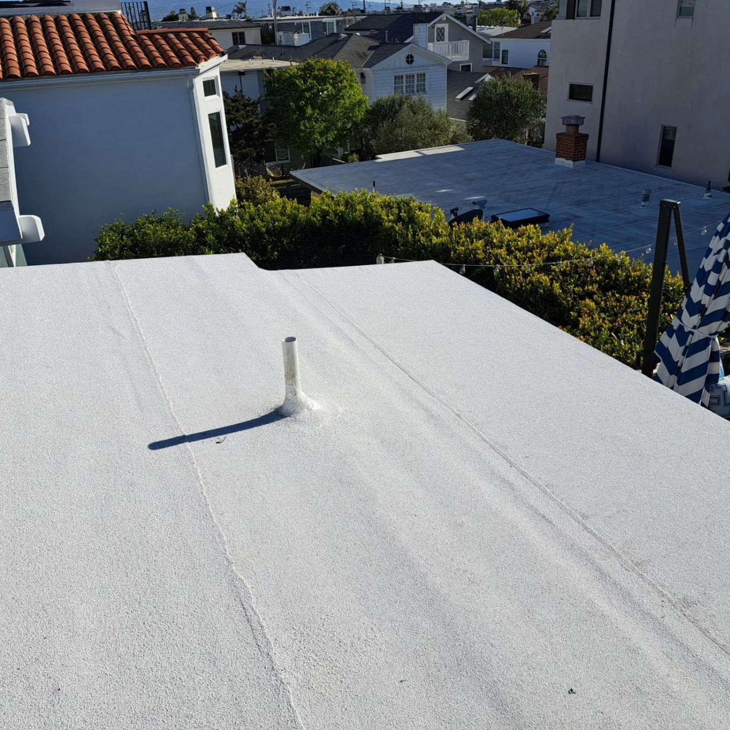 White rooftop terrace with a small vent pipe, hedges, and neighboring houses under bright sun